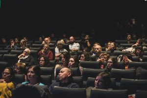 Públic concentrat en una projecció de cinema al Cinema Girona de Barcelona, en una sala fosca. Les persones, majoritàriament adultes, es poden veure atentes i relaxades, amb una postura de concentració mentre miren cap al front. L'atmosfera és tranquil·la, típica d'un cinema d'art i assaig a Barcelona.