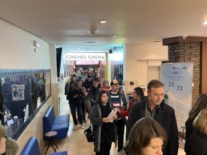 Un grup de persones fent cua dins del Cinema Girona a Barcelona, davant de la seva entrada. La majoria de les persones estan de peu, algunes amb les seves entrades o telèfons mòbils a les mans, mentre que altres conversen. Es veu el cartell de CINEMES GIRONA al fons, amb un ambient modern i acollidor, típic dels cinemes independents de Barcelona.