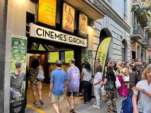 Una multitud a l'exterior del Cinema Girona de Barcelona, esperant per entrar a la sala. Hi ha una gran varietat de persones de diferents edats, algunes fent cua per comprar entrades, d'altres parlant entre elles. Els cartells de pel·lícules a l'exterior mostren títols d'estrena i una gran pancarta publicitària de l'esdeveniment "la GRAN pantalla", destacant l'ambient vibrant de l'oferta cultural de Barcelona.